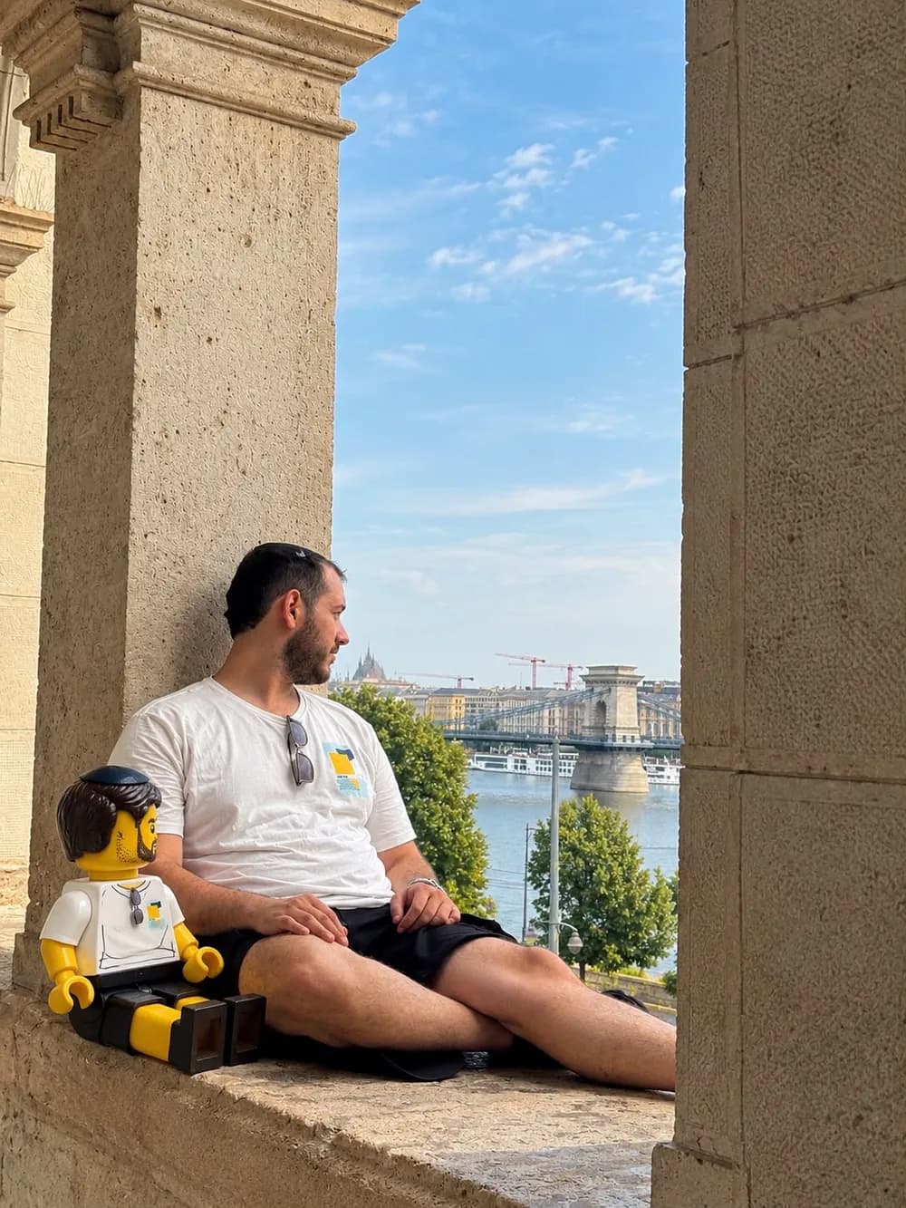 בחור יושב על אדן חלון מול נהר ולידו דמות לגו יושבת עם אותה חולצה ומשקפי שמש